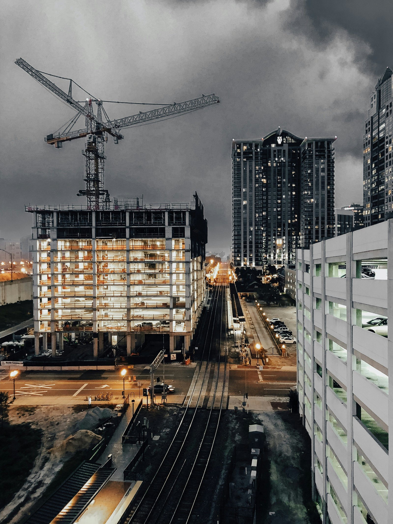 Nighttime cityscape featuring a construction site with a towering crane, an illuminated building under development, railway tracks, and high-rise buildings in the background.